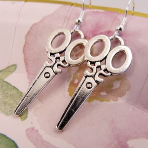 May include: A pair of silver-toned earrings shaped like scissors. The earrings have a detailed design and are a fun and unique accessory.