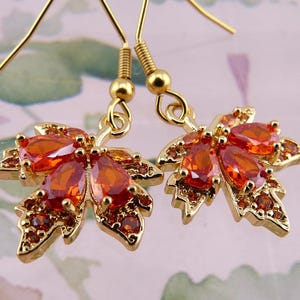 May include: A pair of gold earrings with a maple leaf design. The leaves are adorned with orange and red gemstones.