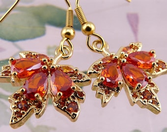 May include: A pair of gold-toned drop earrings shaped like maple leaves. Each earring features multiple pear-shaped and round orange gemstones set into the metal frame, suspended from a gold-toned hook wire for pierced ears.