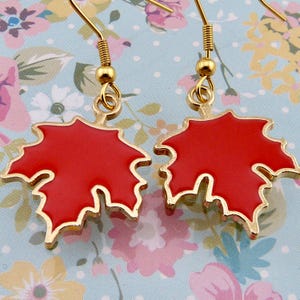 Peut inclure: Une paire de boucles d'oreilles dorées avec des breloques en forme de feuille d'érable rouge. Les feuilles sont bordées d'or et ont une finition brillante.