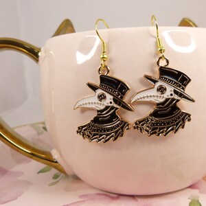 Puede incluir: Un par de pendientes de tono dorado con un dise&ntilde;o de m&eacute;dico de la peste en blanco y negro. Los pendientes cuelgan de una taza rosa con un patr&oacute;n floral.