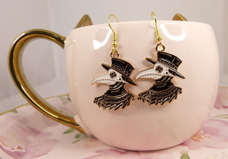 Puede incluir: Un par de pendientes de tono dorado con un dise&ntilde;o de m&eacute;dico de la peste en blanco y negro. Los pendientes cuelgan de una taza rosa con un patr&oacute;n floral.