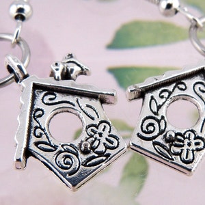 May include: A pair of silver charm earrings featuring a birdhouse design with intricate details. The birdhouses have a small opening and are decorated with swirls and flowers.