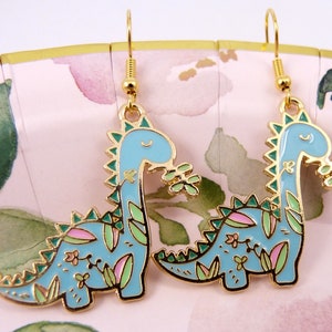 K&ouml;nnte beinhalten: Ein Paar goldfarbene Ohrh&auml;nger mit einem blauen Dinosaurier mit gr&uuml;nen und rosa floralen Details. Die Dinosaurier sind nach rechts gerichtet und haben einen fr&ouml;hlichen Ausdruck.