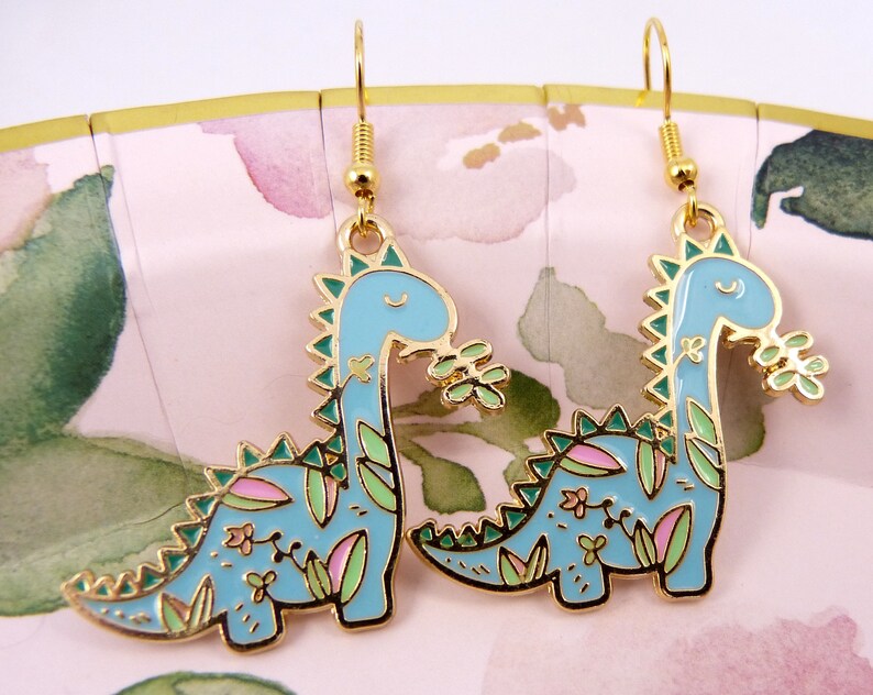K&ouml;nnte beinhalten: Ein Paar goldfarbene Ohrh&auml;nger mit einem blauen Dinosaurier mit gr&uuml;nen und rosa floralen Details. Die Dinosaurier sind nach rechts gerichtet und haben einen fr&ouml;hlichen Ausdruck.