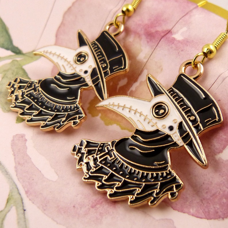 Puede incluir: Un par de pendientes de tono dorado con un dise&ntilde;o de m&eacute;dico de la peste en blanco y negro. Los pendientes tienen un pico largo y delgado, un sombrero negro y un cuello con estampado en blanco y negro.