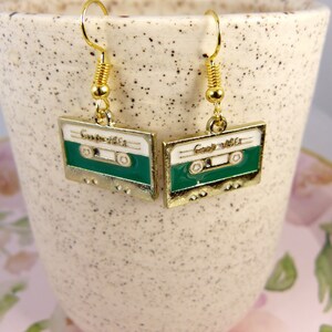 May include: A pair of gold tone earrings with a green and white cassette tape design. The earrings have a spring clasp closure.