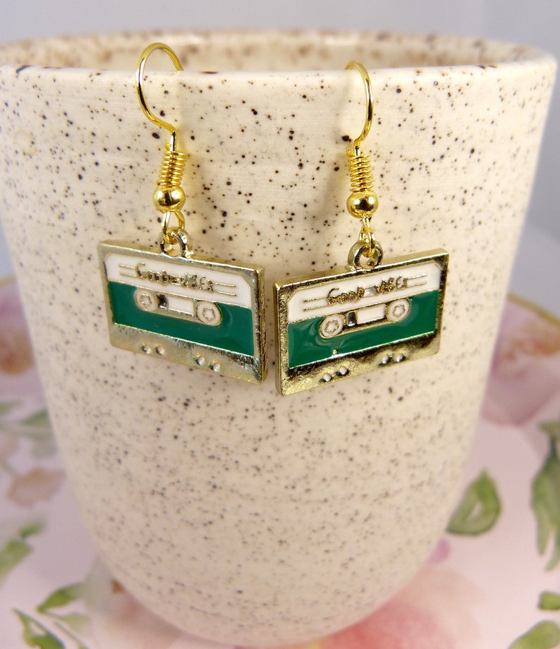 May include: A pair of gold tone earrings with a green and white cassette tape design. The earrings have a spring clasp closure.