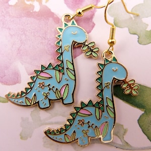 K&ouml;nnte beinhalten: Ein Paar goldfarbene Ohrringe mit blauen Dinosaurier-Anh&auml;ngern mit gr&uuml;nen und rosa floralen Details. Die Dinosaurier halten gr&uuml;ne Bl&auml;tter im Maul.