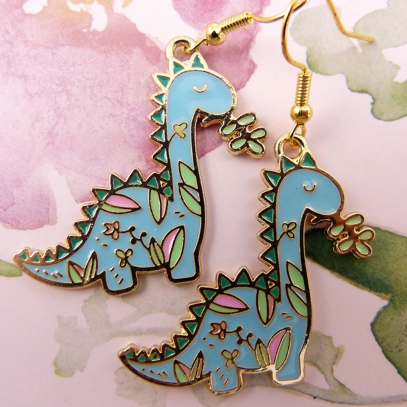 K&ouml;nnte beinhalten: Ein Paar goldfarbene Ohrringe mit blauen Dinosaurier-Anh&auml;ngern mit gr&uuml;nen und rosa floralen Details. Die Dinosaurier halten gr&uuml;ne Bl&auml;tter im Maul.