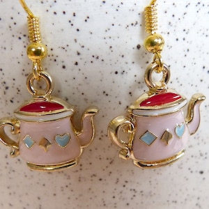 May include: A pair of gold tone earrings featuring a pink teapot design with red, white, and blue accents. The teapots have a diamond, spade, and heart design.