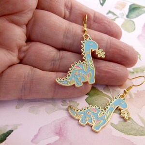 K&ouml;nnte beinhalten: Ein Paar goldfarbene Ohrringe mit einem blauen Dinosaurier mit floralen Details. Der Dinosaurier h&auml;lt eine Blume im Maul.