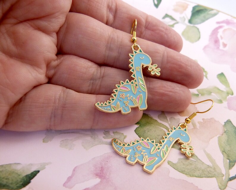 K&ouml;nnte beinhalten: Ein Paar goldfarbene Ohrringe mit einem blauen Dinosaurier mit floralen Details. Der Dinosaurier h&auml;lt eine Blume im Maul.