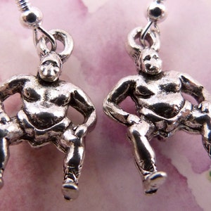 May include: A pair of silver earrings featuring a small, detailed sculpture of a sumo wrestler in a seated pose.