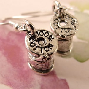 May include: A pair of silver earrings shaped like spools of thread. Each earring has the words "100% Cotton" engraved on the side.