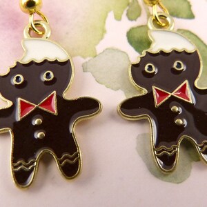 May include: A pair of gingerbread man earrings with a white icing top, red bow tie, and gold accents.