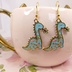 K&ouml;nnte beinhalten: Ein Paar goldene Ohrringe mit einem blauen Dinosaurier mit gr&uuml;nen und rosa floralen Details. Die Dinosaurier sind nach rechts gerichtet und haben ein l&auml;chelndes Gesicht.