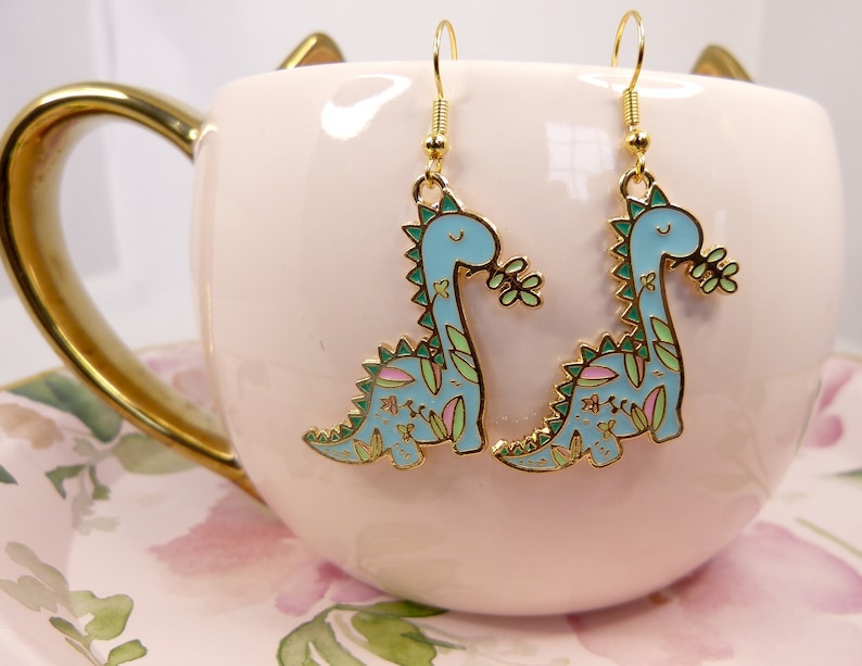 K&ouml;nnte beinhalten: Ein Paar goldene Ohrringe mit einem blauen Dinosaurier mit gr&uuml;nen und rosa floralen Details. Die Dinosaurier sind nach rechts gerichtet und haben ein l&auml;chelndes Gesicht.