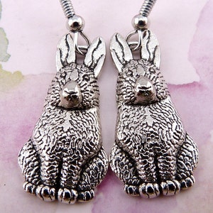 May include: A pair of silver rabbit dangle earrings. The rabbits are sitting with their paws in front of them. The earrings have a textured surface.