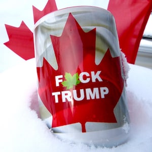 May include: A cylindrical mug with a red maple leaf design and the text "F*CK TRUMP". The mug is set in snow, with a Canadian flag visible in the background.