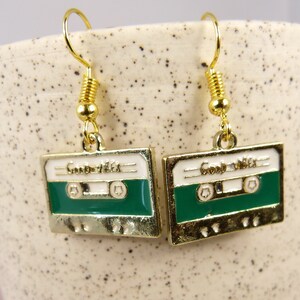 May include: Gold cassette tape earrings with a white background and green tape. The earrings say "Good Vibes" on the tape.