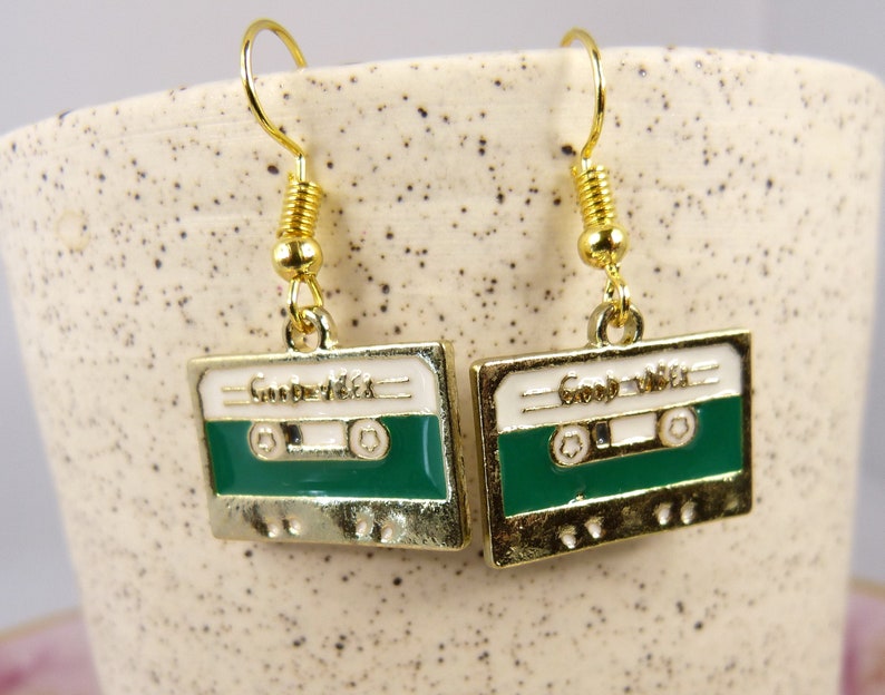 May include: Gold cassette tape earrings with a white background and green tape. The earrings say "Good Vibes" on the tape.