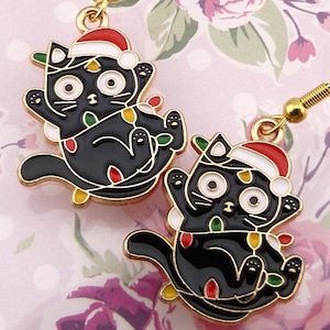 May include: A pair of gold-coloured cat earrings. Each black cat is wearing a Santa hat and wrapped in colourful Christmas lights. The earrings have a festive, holiday theme.