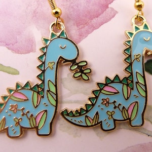 K&ouml;nnte beinhalten: Ein Paar goldfarbene Ohrringe mit einem blauen Dinosaurier-Design mit gr&uuml;nen und rosa floralen Akzenten.
