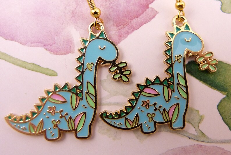 K&ouml;nnte beinhalten: Ein Paar goldfarbene Ohrringe mit einem blauen Dinosaurier-Design mit gr&uuml;nen und rosa floralen Akzenten.
