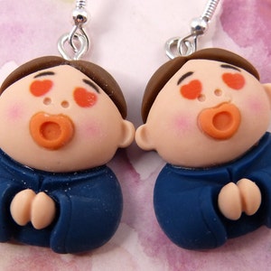 May include: A pair of handmade earrings featuring two cartoon figures with brown hair, rosy cheeks, and orange lips. The figures are wearing blue outfits and have heart-shaped eyes. The earrings have silver-colored hooks.