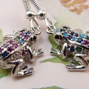 May include: A pair of silver frog earrings with colorful gemstones. The frogs are detailed with sparkling blue, green, pink, and turquoise stones.