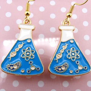 Peut inclure: Une paire de boucles d'oreilles dorées en forme de béchers avec un liquide bleu et des symboles scientifiques, notamment une chaîne d'ADN, un atome, une bactérie et une molécule.