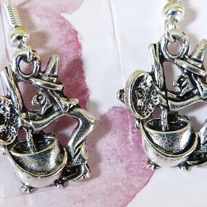 May include: A pair of silver earrings featuring a witch stirring a cauldron. The witch is depicted in a stylised, whimsical design with a pointed hat and a long, flowing robe. The cauldron is filled with a bubbling liquid, and the witch is holding a ladle in her hand.