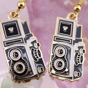 May include: A pair of gold-tone earrings featuring a black and white enamel camera design. The camera has two lenses and a small viewfinder on top.