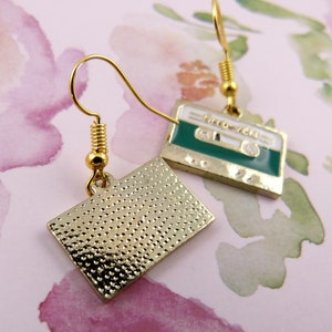 May include: A pair of gold tone earrings with a cassette tape design. One earring has a green cassette tape with the text "GROOVE TAPE" on it. The other earring has a gold tone cassette tape with a textured surface.