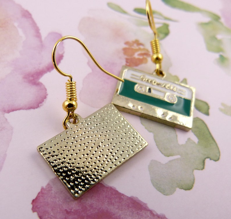 May include: A pair of gold tone earrings with a cassette tape design. One earring has a green cassette tape with the text "GROOVE TAPE" on it. The other earring has a gold tone cassette tape with a textured surface.