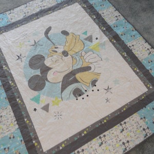 May include: A white, blue, and gray baby quilt with a Mickey Mouse and Pluto design. The quilt features a blue circle with Mickey Mouse and Pluto in the center. The background of the quilt is white with a light blue and gray geometric pattern.