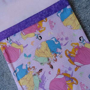 Pu&ograve; includere: Un tessuto rosa con un motivo a pois bianchi che presenta diverse principesse Disney nei loro abiti iconici. Il tessuto ha un bordo di nastro viola con un motivo floreale.