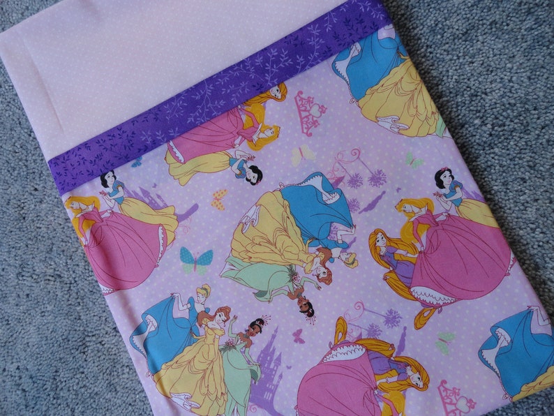 Pu&ograve; includere: Un tessuto rosa con un motivo a pois bianchi che presenta diverse principesse Disney nei loro abiti iconici. Il tessuto ha un bordo di nastro viola con un motivo floreale.