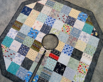 May include: A gray and white patchwork Christmas tree skirt with a variety of holiday-themed fabrics. The skirt has a hole in the center for a tree stand.
