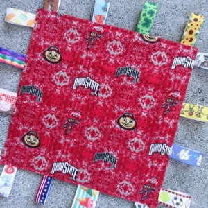 May include: Red baby blanket with a variety of colorful ribbon tags. The blanket features a repeating pattern of the Ohio State logo and a cartoon mascot. The tags include a rainbow, a sunflower, a teddy bear, a soccer ball, a tooth, and more.
