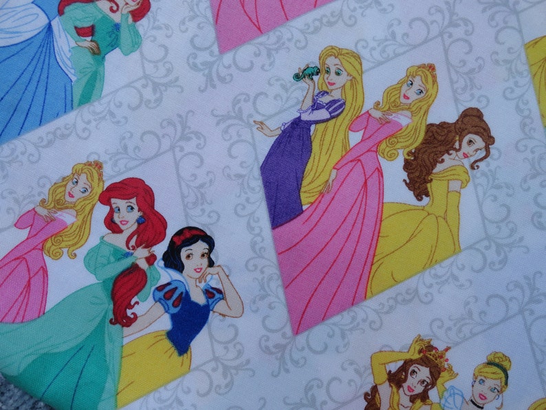 Pu&ograve; includere: Un tessuto bianco con un motivo ripetuto di principesse Disney in abiti colorati. Le principesse includono Ariel, Belle, Cenerentola, Biancaneve e Rapunzel.