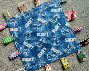 May include: A blue baby blanket with a white "Lions" pattern and multiple colorful fabric tags. The tags feature various designs, including a rainbow, a watermelon, a cow, and a cartoon character.