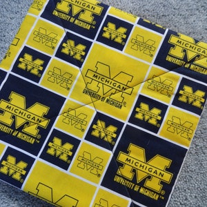 May include: A yellow and blue fabric with the University of Michigan logo repeated in a checkered pattern. The logo is a block "M" with the words "Michigan" and "University of Michigan" underneath.