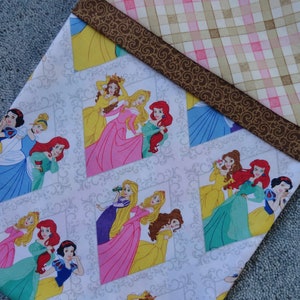 Pu&ograve; includere: Tessuto bianco con un motivo ripetuto di principesse Disney in cornici. Le principesse sono Biancaneve, Cenerentola, Ariel, Belle, Aurora e Rapunzel. Il tessuto ha un bordo a quadri marrone e rosa.