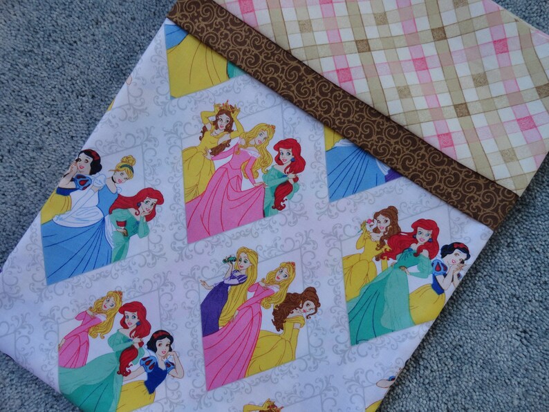 Pu&ograve; includere: Tessuto bianco con un motivo ripetuto di principesse Disney in cornici. Le principesse sono Biancaneve, Cenerentola, Ariel, Belle, Aurora e Rapunzel. Il tessuto ha un bordo a quadri marrone e rosa.