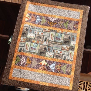 May include: A brown and orange quilted throw blanket with a woodland theme. The blanket features a variety of animals, plants, and other nature-inspired designs.