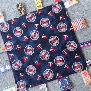 May include: A blue baby blanket with a repeating pattern of red "Twins" logos. The blanket has multiple fabric tags attached to the edges, including a rainbow, a piano, a hamburger, and a fruit pattern.
