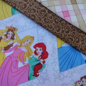 Pu&ograve; includere: Un tessuto bianco con un motivo a spirale marrone e un motivo a quadri rosa e beige. Il tessuto presenta un'immagine stampata di quattro principesse Disney: Belle, Aurora, Ariel e Cenerentola. Ogni principessa indossa un abito di colore diverso e ha un'acconciatura diversa.