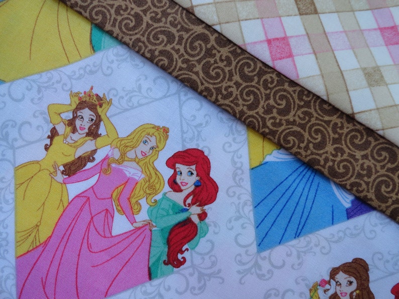 Pu&ograve; includere: Un tessuto bianco con un motivo a spirale marrone e un motivo a quadri rosa e beige. Il tessuto presenta un'immagine stampata di quattro principesse Disney: Belle, Aurora, Ariel e Cenerentola. Ogni principessa indossa un abito di colore diverso e ha un'acconciatura diversa.